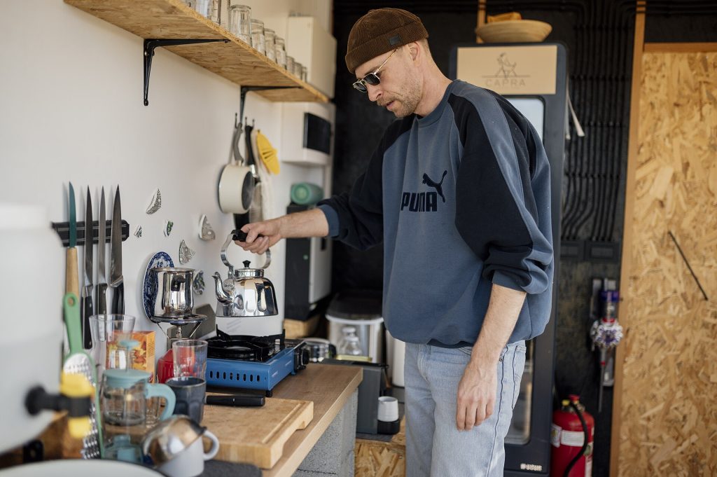 Man makes coffee in a small kitchen with wooden shelves and various kitchen tools